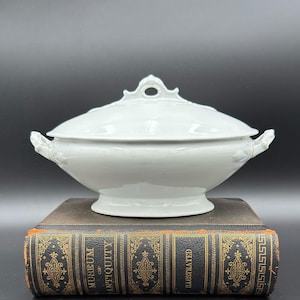 May include: A white ceramic tureen with a matching lid featuring a central handle with a circular opening and two side handles. The tureen has a smooth, glossy finish and sits on a pedestal base, showcasing a clean, rounded, and symmetrical design.