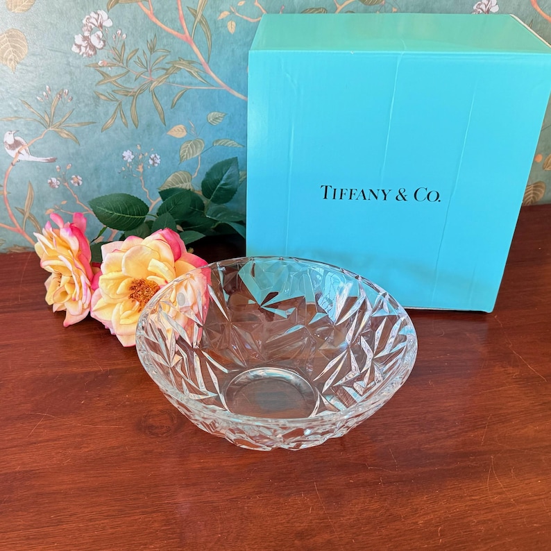 May include: A clear, faceted glass bowl sits in front of a Tiffany & Co. box. The box is a bright turquoise color, and the text "Tiffany & Co." is printed on the front. Pink and yellow roses are to the left of the bowl.