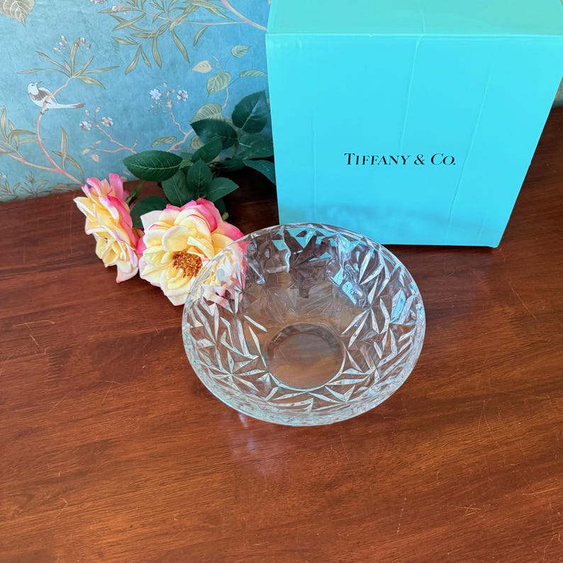 May include: A clear, cut-glass bowl with a geometric pattern sits on a wooden surface. A Tiffany & Co. box is in the background, along with pink and yellow roses. The bowl is round and has a textured surface.