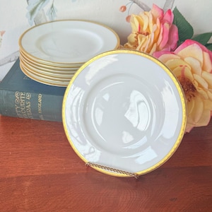 Op de afbeelding: Witte porseleinen borden met een gouden rand. Een stapel borden is zichtbaar, met één bord op de voorgrond op een gouden standaard. De borden staan op een houten oppervlak. De borden maken deel uit van een servies.