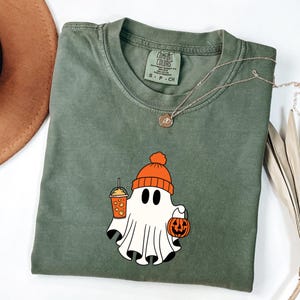 May include: Olive green Comfort Colors t-shirt featuring a cartoon ghost wearing an orange beanie and holding a pumpkin and a drink. A silver necklace with a pendant is draped across the shirt.