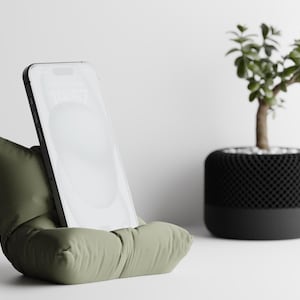 May include: An olive green phone stand with a pillow-like design, holding a smartphone. A small potted plant with a black and gray pot is in the background. The phone displays the time.