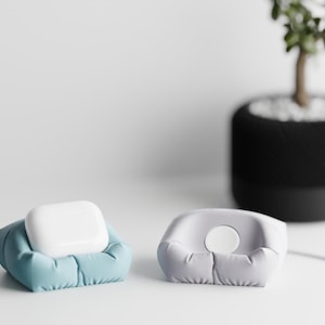 May include: Two small, pillow-like holders, one blue and one gray, designed to cradle small electronic devices. A white device rests in the blue holder. A small potted plant is in the background.