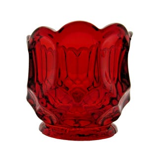 May include: A ruby red glass votive candle holder with a scalloped rim and a detailed, textured design. The base is round and slightly raised. The candle holder is made of transparent glass.