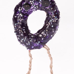 May include: A purple donut with iridescent sprinkles and two black buttons for eyes, standing on two twisted brown legs.