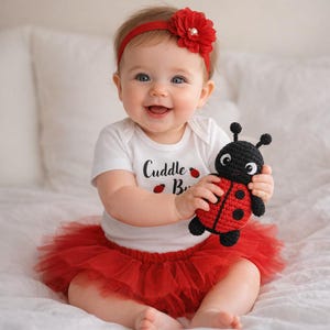 Puede incluir: Un bebé sonriente con una diadema roja con una flor, un body blanco con el texto "Cuddle Bug" y un tutú rojo. El bebé sostiene un juguete de mariquita de ganchillo negro y rojo.
