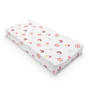 Könnte beinhalten: Weißer Spannbezug für Wickelauflagen mit einem sich wiederholenden Muster aus rosa und gelben Blumen, Regenbogen und Wolken. Der Bezug ist für einen Wickeltisch für Babys konzipiert und besteht aus weichem Stoff.