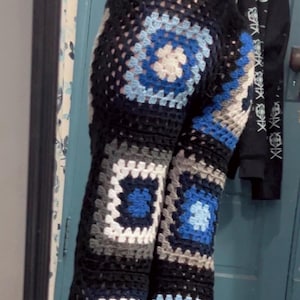 Puede incluir: Pantalones de ganchillo con un patrón de cuadrados granny en tonos negro, blanco, azul claro y azul oscuro. Los pantalones están confeccionados con múltiples motivos cuadrados unidos, creando un diseño texturizado y calado de corte largo.