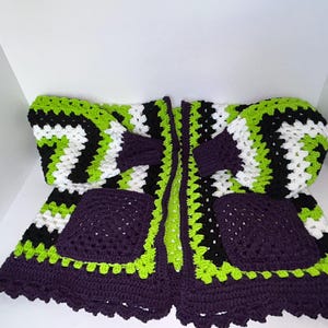 May include: A handmade crochet cardigan in a striking colour scheme. The open-front design features a combination of black, white, and neon green, with deep purple accents on the pockets and trim. The cardigan has long sleeves and two front pockets.