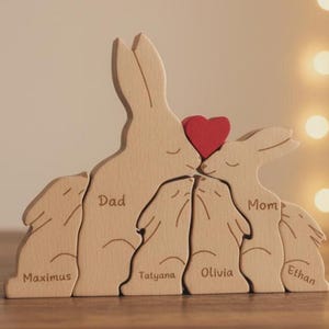 May include: Wooden family bunny puzzle with a red heart. The puzzle features a dad, mom, and three children named Maximus, Tatyana, Olivia, and Ethan. The bunnies are made of light-colored wood.