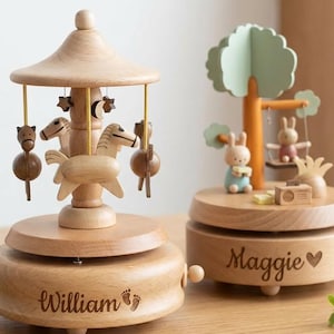 May include: Two wooden music boxes. One is a carousel with horses and stars, the other features a tree with bunnies on a swing. The carousel base is engraved with "William" and footprints. The other base is engraved with "Maggie" and a heart.