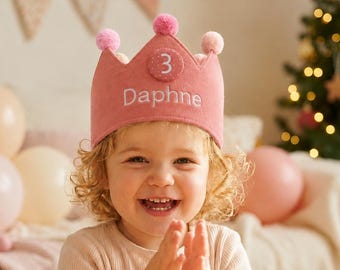 May include: A dusty pink felt crown featuring the name Daphne embroidered in white thread and a circular patch with the number 3. The crown is adorned with small pink pom-poms along the top edge, creating a festive accessory for a birthday celebration.
