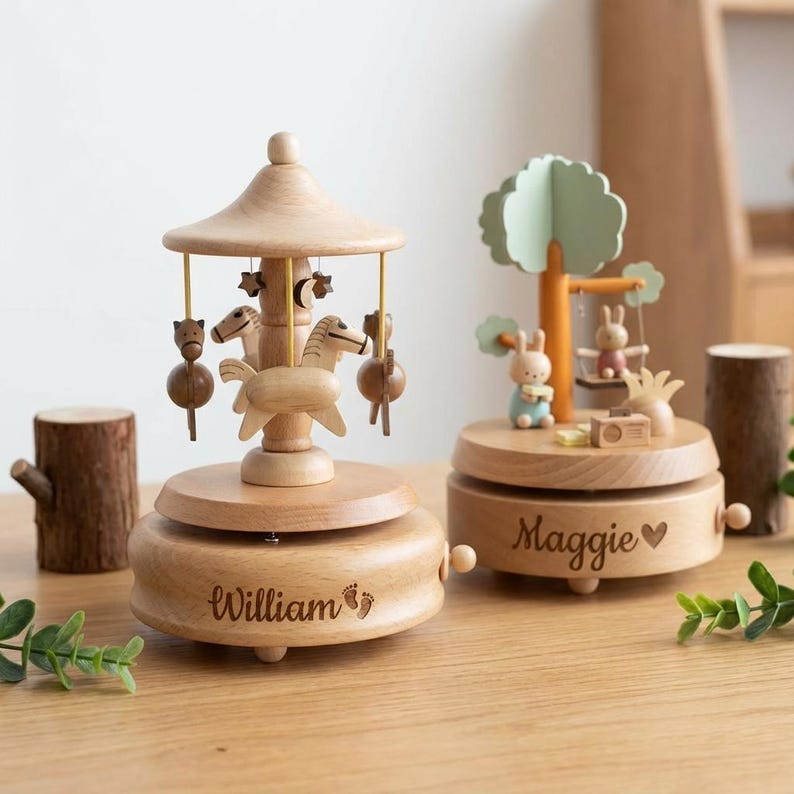 May include: Two wooden music boxes. One is a carousel with horses and stars, the other features a tree with bunnies on a swing. The carousel base is engraved with "William" and footprints. The other base is engraved with "Maggie" and a heart.