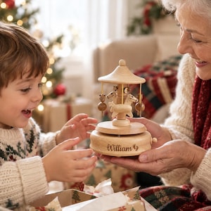 May include: A light-colored wooden carousel music box featuring carved horses suspended from a central canopy. The circular base is engraved with the name William and a small footprint icon. The structure is made of smooth, polished wood.