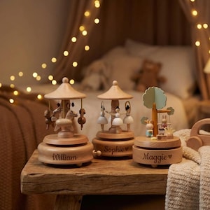 May include: Three wooden music boxes with personalized names. One features horses, another ballerinas, and the third has a tree with bunnies on a swing. The boxes are light brown and sit on a wooden table.