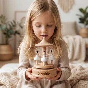 May include: A wooden carousel music box featuring three ballerina figurines in white dresses. The music box has a light brown finish and the name "Sophie" is engraved on the base. The carousel is held by a young girl.