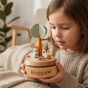 May include: A wooden music box featuring a tree with two bunny figurines, one on a swing. The base of the box is engraved with the name "Maggie". The music box has a light wood finish and includes small decorative elements.