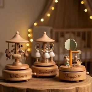 May include: Three wooden music boxes with carousel and woodland themes. The first features horses, the second ballerinas, and the third bunnies. Each box has a name engraved on it: William, Sophie, and Maggie. The boxes are made of light-colored wood.