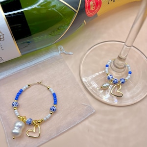 Puede incluir: Una botella de champán Moët con dos colgantes para copas de vino. Los colgantes son aros dorados con cuentas azules y blancas, colgantes de corazón dorados y una perla en forma de gota. Un colgante está en una copa de vino.