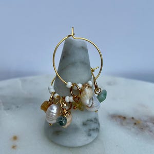 May include: Gold hoop earrings featuring charms like pearls, seashells, and gemstones. The earrings are presented on a marble stand, highlighting their intricate design and coastal theme. The earrings are a beautiful accessory.