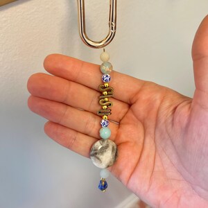 May include: A beaded keychain with a gold-colored clasp. The keychain features a series of beads in various colors, including blue, white, and gray, with the word "SHSH" in gold-colored letters. The keychain is held by a hand.