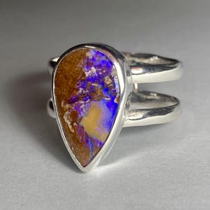 May include: A sterling silver ring featuring a pear-shaped opal. The opal displays a mix of brown, yellow, purple, and blue colors. The ring has a double-band design.
