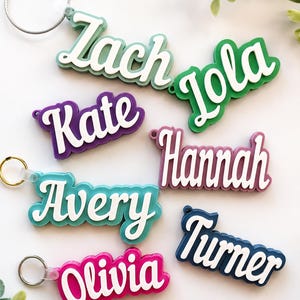 May include: A collection of colorful name keychains. Each keychain features a different name in a white, cursive font, set against a vibrant background. Colors include green, purple, blue, pink, and navy. Each keychain has a metal ring for attachment.