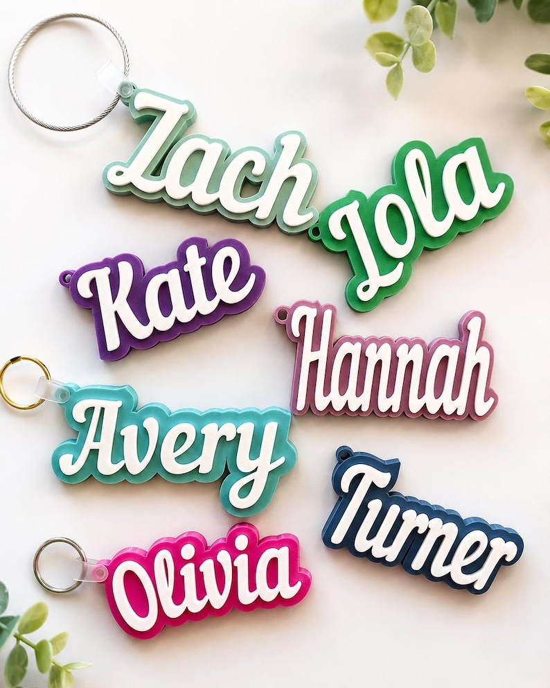 May include: A collection of colorful name keychains. Each keychain features a different name in a white, cursive font, set against a vibrant background. Colors include green, purple, blue, pink, and navy. Each keychain has a metal ring for attachment.