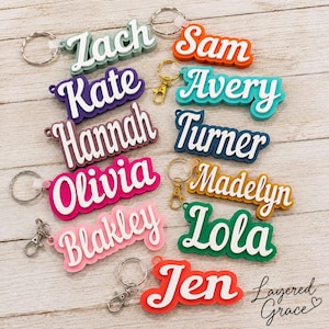 May include: A collection of colorful name keychains in various colors, including green, orange, pink, and blue. Each keychain features a different name in a stylish, layered design with a metal keyring and clasp.