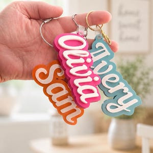 May include: Three personalized name keychains in orange, pink, and blue. Each keychain features a name in a white, layered, cursive font. The keychains are attached to metal rings and a wire loop.