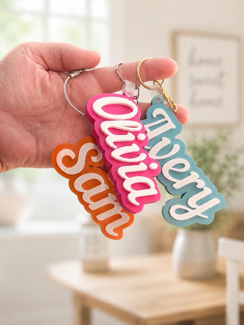 May include: Three personalized name keychains in orange, pink, and blue. Each keychain features a name in a white, layered, cursive font. The keychains are attached to metal rings and a wire loop.