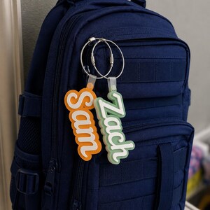 May include: A navy blue backpack with two personalized name tags attached. One tag is orange and says "Sam", the other is green and says "Zach". The tags are connected to the backpack with silver key rings. The backpack has multiple pockets and straps.