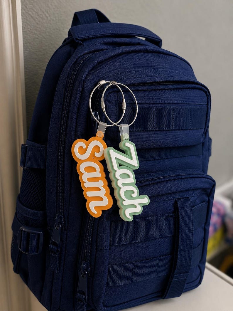 May include: A navy blue backpack with two personalized name tags attached. One tag is orange and says "Sam", the other is green and says "Zach". The tags are connected to the backpack with silver key rings. The backpack has multiple pockets and straps.