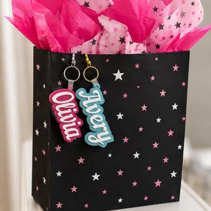 May include: A black gift bag with a star pattern, filled with bright pink tissue paper. Two keychains with the names "Olivia" and "Avery" hang from the bag. The keychains have a white outline and are in pink and blue.