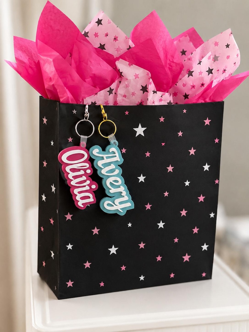 May include: A black gift bag with a star pattern, filled with bright pink tissue paper. Two keychains with the names "Olivia" and "Avery" hang from the bag. The keychains have a white outline and are in pink and blue.