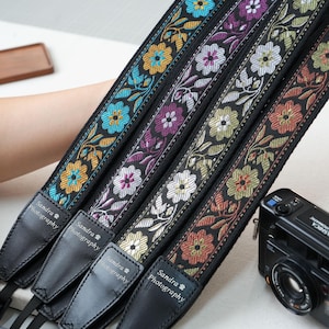 May include: A collection of black camera straps with floral embroidery in various colors, including blue, purple, and orange. Each strap has a leather end with the text "Sandra Photography". A black camera is visible in the background.