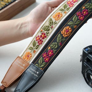 以下が含まれることがあります： 花柄刺繍のカメラストラップ2本。1本はオレンジとピンクの花柄の黒、もう1本はオレンジと赤の花柄のクリーム色。どちらも「Sandra Photography」の文字が入った革製の端が付いています。背景には黒いカメラがあります。