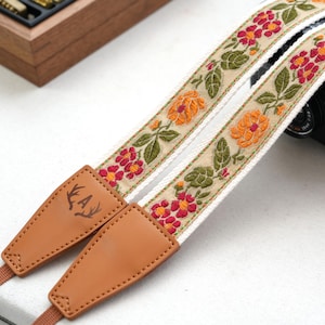 May include: Camera strap with a floral embroidered design. The strap features a cream-colored base with red and orange flowers and green leaves. The ends are brown leather with an engraved design.