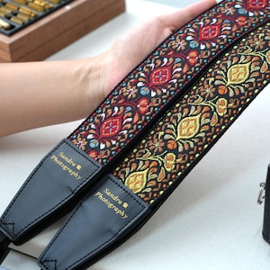 Può includere: Due cinghie decorative per fotocamera con intricati motivi floreali in rosso, arancione, giallo e blu su sfondo nero. Le cinghie hanno estremità in pelle nera con la scritta "Sandra Photography". Una fotocamera nera è visibile sullo sfondo.