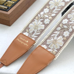 Könnte beinhalten: Ein Kameragurt mit floralem Stickmuster in Weiß und Gold auf taupefarbenem Grund. Der Gurt hat braune Lederenden mit dem Text "Sandra Photography". Die Kanten sind mit einer cremefarbenen Bordüre versehen.
