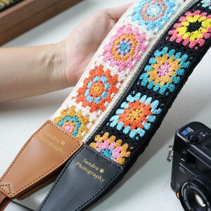 May include: Two crochet camera straps with a floral pattern in orange, yellow, pink, and blue. One strap is black, the other is white. The straps have leather ends with the text "Sandra Photography".