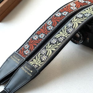 Può includere: Due cinghie per fotocamera con motivi floreali rossi, bianchi e verdi su sfondo nero. Le cinghie hanno estremità in pelle nera con la scritta "Sandra Photography". Una fotocamera nera è parzialmente visibile sullo sfondo.