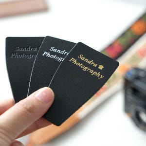 May include: Black business cards with the text "Sandra Photography" in gold lettering. The cards are held in front of a blurred background with a camera and patterned straps.