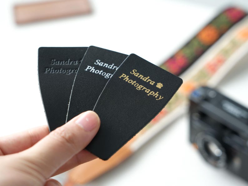 May include: Black business cards with the text "Sandra Photography" in gold lettering. The cards are held in front of a blurred background with a camera and patterned straps.