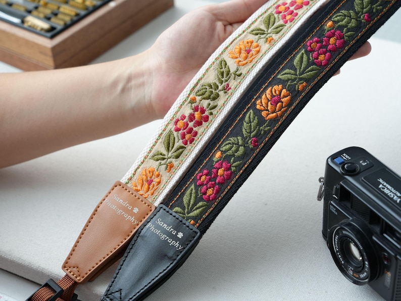May include: Two embroidered camera straps with floral designs. One strap is black with orange and pink flowers, the other is cream with orange and red flowers. Both straps have leather ends with the text "Sandra Photography". A black camera is in the background.