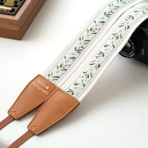 May include: A camera strap with a cream-colored background and a floral pattern in green and brown. The strap has brown leather ends with the text "Sandra Photography" in gold. A black camera is partially visible.