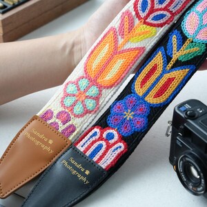 May include: Two embroidered camera straps with floral patterns. One is white with orange, pink, and blue flowers. The other is black with blue, red, and yellow flowers. Both straps have "Sandra Photography" on the leather ends.