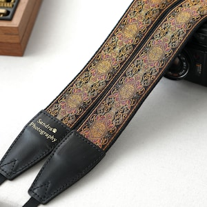 Può includere: Una tracolla per fotocamera con un motivo decorativo in oro, rosa e nero. La tracolla ha estremità in pelle nera con la scritta "Sandra Photography". Una fotocamera nera è parzialmente visibile sullo sfondo.