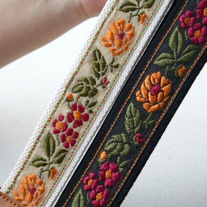 May include: Two embroidered camera straps with floral patterns. One strap is black with orange and pink flowers, the other is off-white with orange and red flowers. Both straps have green leaves and brown leather ends.