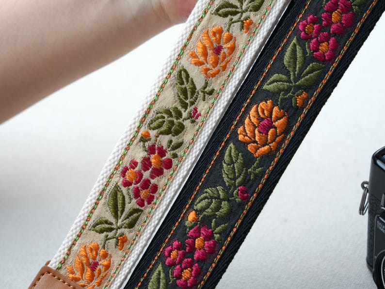 May include: Two embroidered camera straps with floral patterns. One strap is black with orange and pink flowers, the other is off-white with orange and red flowers. Both straps have green leaves and brown leather ends.